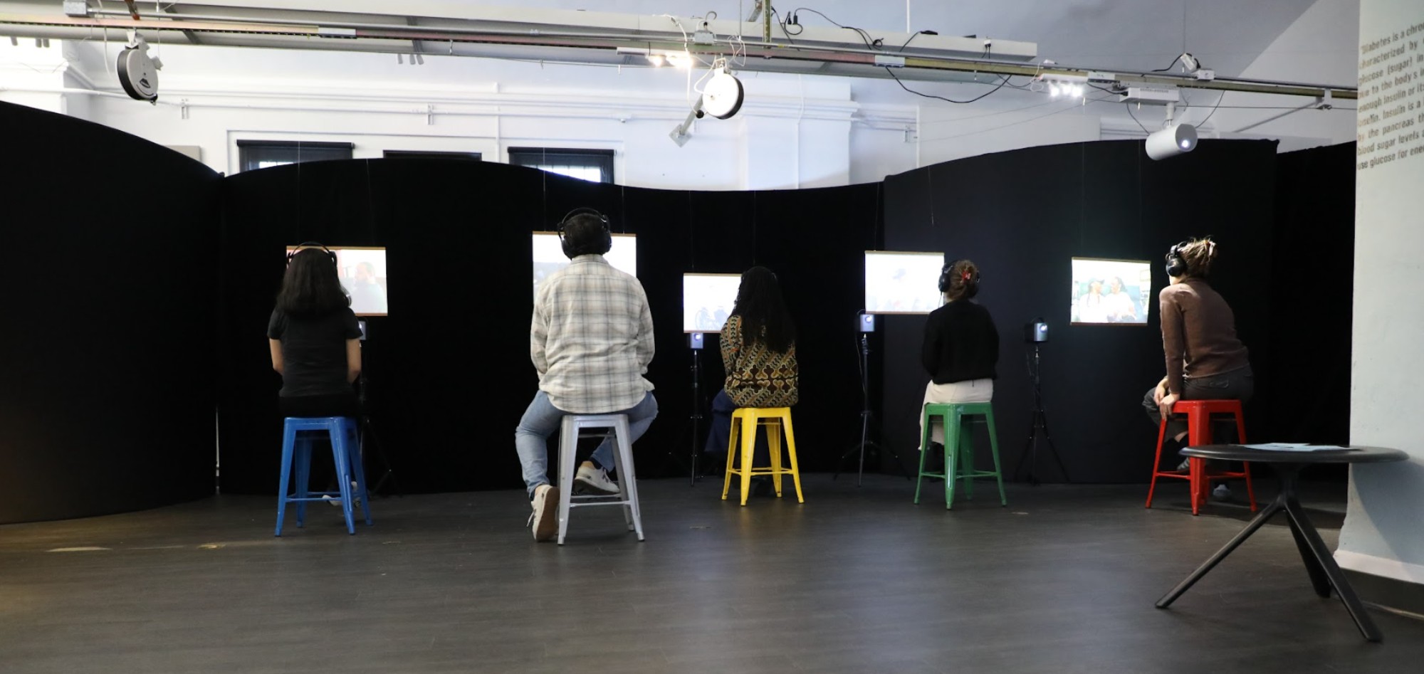 Five exhibit visitors sit on 5 different colored stools (from left to right: blue, silver, yellow, green, red) and look at screens with headphones on. The photo was taken of them from behind, so only the backs of their bodies are visible. There is a black fabric background behind the individual video screens.
